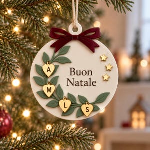 Könnte beinhalten: Ein runder, weißer Weihnachtsanhänger mit einer dunkelroten Schleife oben und einem grünen Blattkranz-Design. Vier goldene herzförmige Anhänger mit den Buchstaben A, M, L und S sind befestigt. Der Text Buon Natale ist in Dunkelbraun aufgedruckt.