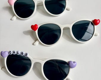 May include: Three pairs of white-framed sunglasses with dark lenses. Two pairs have heart-shaped accents, one pink and one red. The third pair has a purple heart and the name "CHLOE" spelled out in white letter beads.