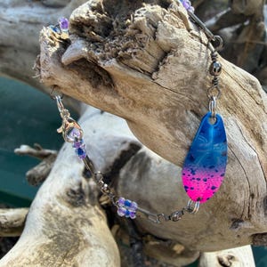 Może przedstawiać: Srebrna bransoletka z niebiesko-różowym gradientowym urokiem. Urok ma kształt łzy i ma teksturowaną powierzchnię. Bransoletka posiada małe fioletowe i niebieskie koraliki i jest prezentowana na kawałku drewna wyrzuconego na brzeg.