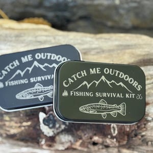 Puede incluir: Dos latas rectangulares de metal con esquinas redondeadas. Las latas son de color gris oscuro y verde oliva, cada una con texto blanco que dice "Catch Me Outdoors Fishing Survival Kit" y una ilustración de un pez. Las latas están sobre un trozo de madera flotante.