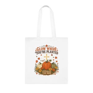 Könnte beinhalten: Eine weiße Tragetasche mit einem Grafikdruck, der den Text GLOW WHERE YOU'RE PLANTED über einem Kreuz, drei Kürbissen in Orange, Weiß und Grün, Herbstblättern und einem Schild mit der Aufschrift PUMPKIN PATCH sowie einem kleinen roten Herz zeigt.