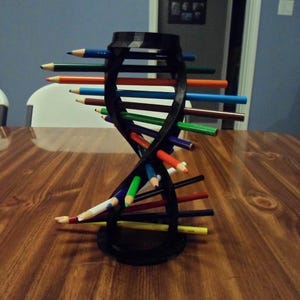 Puede incluir: Un portalápices negro con forma de ADN, lleno de lápices de colores. El soporte está sobre una mesa de madera. Los lápices están dispuestos en espiral, con una variedad de colores visibles.