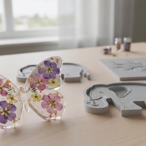 May include: A clear resin butterfly with embedded purple, pink, and yellow flowers. Beside it are gray molds in the shapes of a butterfly, an elephant, and a square with a tree design. The items are on a light wood surface.