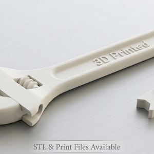 Può includere: Una chiave inglese regolabile stampata in 3D e un piccolo pezzo separato, entrambi di colore beige chiaro. La chiave ha la scritta "3D Printed" sul manico. Gli oggetti sono su una superficie grigio chiaro.