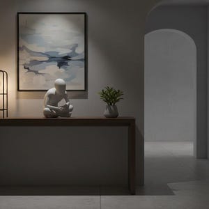 May include: A modern interior scene featuring a dark wood console table. On the table are a black metal cage lamp with a lit bulb, a white sculpture of a person in a seated pose, and a small vase with greenery. A framed abstract painting hangs on the wall.