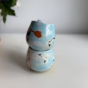 May include: Two stacked, handmade ceramic mugs. The mugs are light blue with hand-painted designs of seagulls, a witch on a broom, and clouds. The mugs have a rounded shape and a small handle.