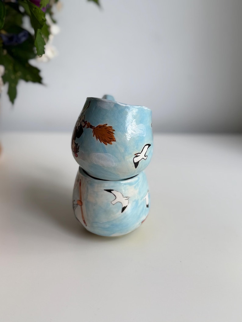 May include: Two stacked, handmade ceramic mugs. The mugs are light blue with hand-painted designs of seagulls, a witch on a broom, and clouds. The mugs have a rounded shape and a small handle.
