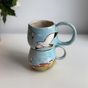 May include: Two ceramic mugs stacked, featuring a hand-painted design. The mugs are light blue with images of seagulls and a coastal town. The handles are a matching light blue. The mugs are on a white surface.