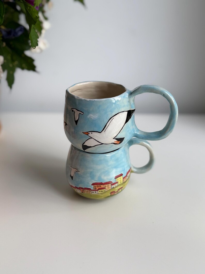 May include: Two ceramic mugs stacked, featuring a hand-painted design. The mugs are light blue with images of seagulls and a coastal town. The handles are a matching light blue. The mugs are on a white surface.