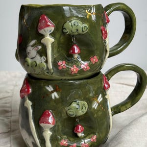 May include: Two stacked, handcrafted ceramic mugs with a whimsical forest theme. The mugs are a deep green with raised relief details of mushrooms, a rabbit, clouds, dragonflies, and pink flowers. Each mug has a large, curved handle.