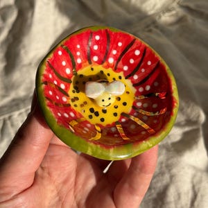 May include: A small, hand-painted ceramic dish with a whimsical design. The dish features a red interior with white polka dots and a yellow centre with black spots. A small, white bow is in the centre. The rim is green.