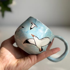 May include: A ceramic mug with a light blue exterior, featuring hand-painted white seagulls in flight against a sky-blue background. The mug has a rounded handle and is held in a hand.