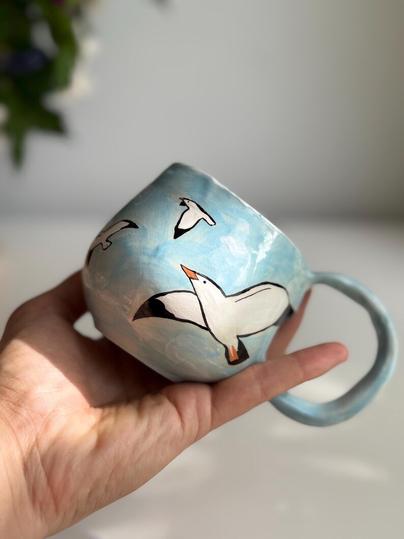 May include: A ceramic mug with a light blue exterior, featuring hand-painted white seagulls in flight against a sky-blue background. The mug has a rounded handle and is held in a hand.