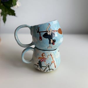 May include: Two light blue ceramic mugs stacked on top of each other. The top mug features a cartoon of a witch on a broom, and the bottom mug has a cartoon of a person riding a bicycle. Both mugs have a light blue background.