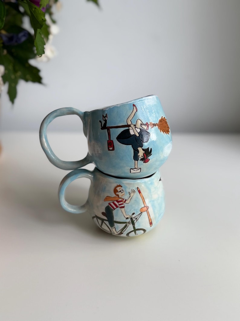 May include: Two light blue ceramic mugs stacked on top of each other. The top mug features a cartoon of a witch on a broom, and the bottom mug has a cartoon of a person riding a bicycle. Both mugs have a light blue background.
