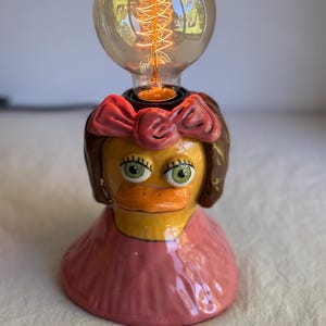 May include: A ceramic table lamp shaped like a duck wearing a pink dress and a matching pink bow in its brown hair. The duck has a yellow face, orange beak, and green eyes. A round, clear glass Edison-style light bulb sits atop the duck's head.