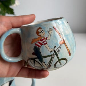 May include: A light blue ceramic mug with a whimsical illustration of a person riding a bicycle with a propeller. The person wears glasses, a red and white striped shirt, and a scarf. The mug has a curved handle.