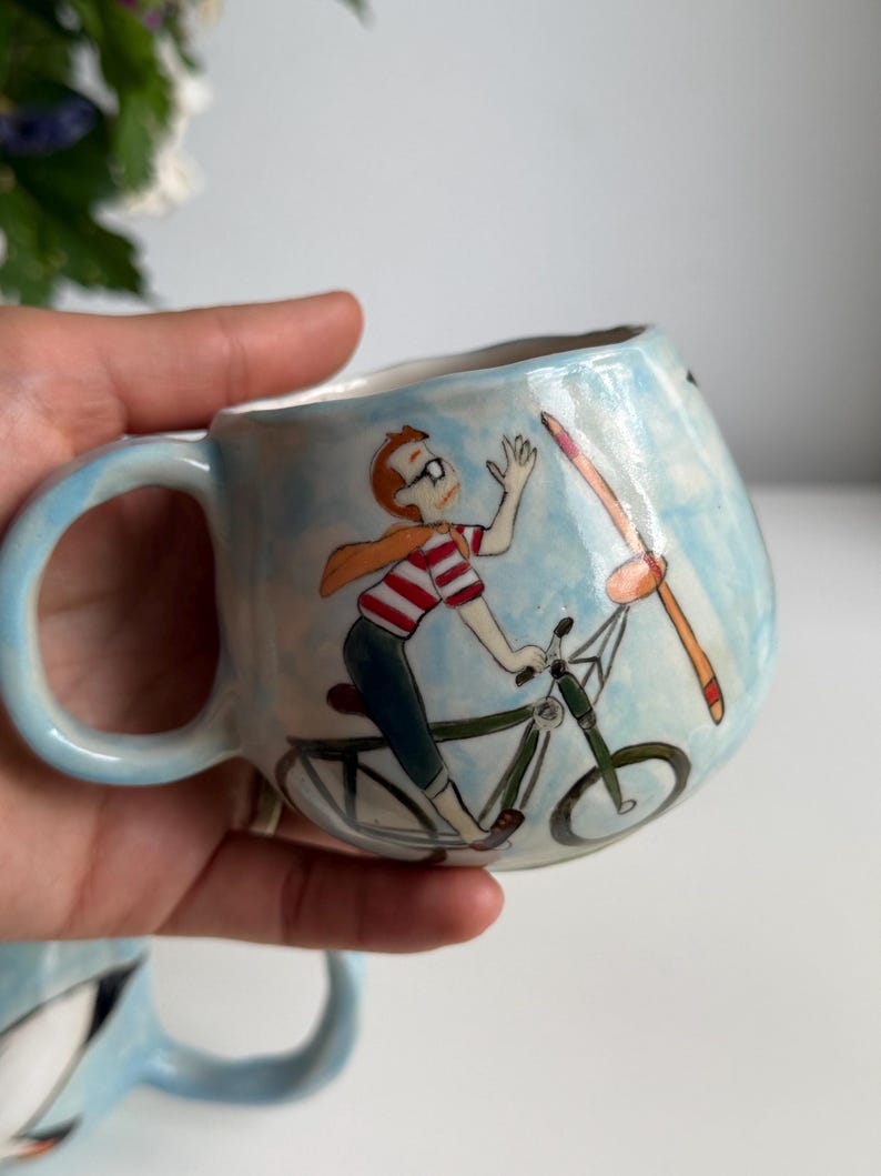 May include: A light blue ceramic mug with a whimsical illustration of a person riding a bicycle with a propeller. The person wears glasses, a red and white striped shirt, and a scarf. The mug has a curved handle.