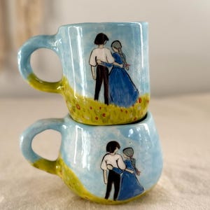 May include: Two handmade ceramic mugs stacked vertically, each featuring a hand-painted scene of two figures standing in a field under a blue sky. The figures are depicted from behind, with one wearing a white shirt and dark trousers, and the other in a blue dress.