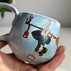 May include: A light blue ceramic mug with a whimsical illustration. The design features a cartoon girl on a broom, a black cat, and a red bag. The mug has a curved handle and a rounded shape.