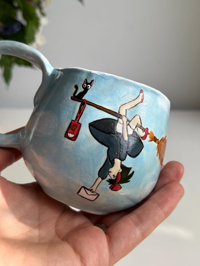 May include: A light blue ceramic mug with a whimsical illustration. The design features a cartoon girl on a broom, a black cat, and a red bag. The mug has a curved handle and a rounded shape.