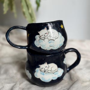 Può includere: Due tazze in ceramica impilate con una glassa blu scuro. Ogni tazza presenta un gatto bianco addormentato su un design a nuvola con stelle gialle. Le tazze hanno manici curvi e una finitura lucida.