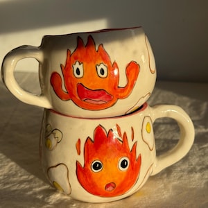 May include: Two off-white ceramic mugs stacked vertically, each featuring a hand-painted orange and red flame character with wide eyes and a mouth. The mugs are decorated with small illustrations of fried eggs and have a glossy finish.