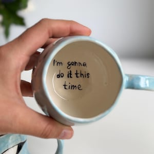 May include: A light blue ceramic mug with a handle, held in a hand. The interior of the mug is off-white, with the handwritten phrase "I'm gonna do it this time" in black ink. The mug has a rounded shape.