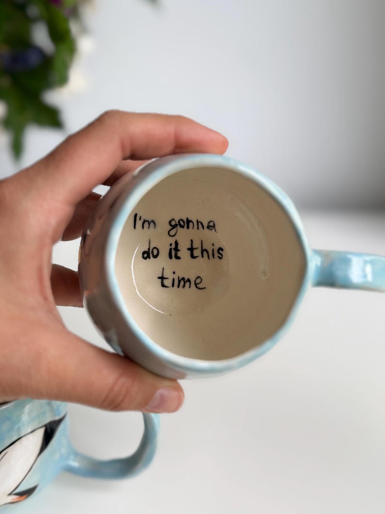 May include: A light blue ceramic mug with a handle, held in a hand. The interior of the mug is off-white, with the handwritten phrase "I'm gonna do it this time" in black ink. The mug has a rounded shape.