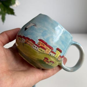 May include: A hand-painted ceramic mug with a whimsical village scene. The mug features a light blue sky, green hills, and colorful houses with red roofs. A white seagull flies across the sky. The mug has a curved handle.