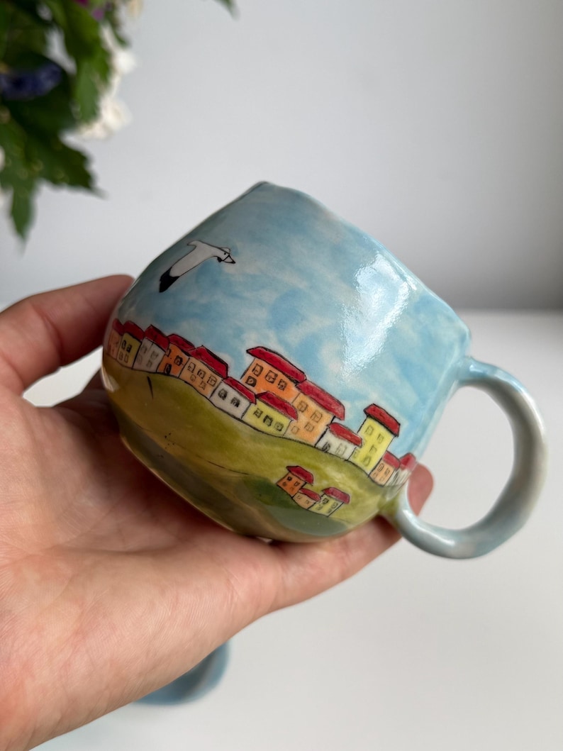 May include: A hand-painted ceramic mug with a whimsical village scene. The mug features a light blue sky, green hills, and colorful houses with red roofs. A white seagull flies across the sky. The mug has a curved handle.