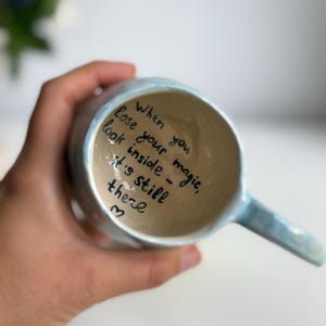 May include: A light blue ceramic mug with a handle, held in a hand. The inside of the mug has handwritten text that reads: "When you lose your magic, look inside - it's still there" with a heart symbol.