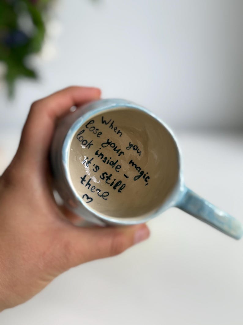 May include: A light blue ceramic mug with a handle, held in a hand. The inside of the mug has handwritten text that reads: "When you lose your magic, look inside - it's still there" with a heart symbol.