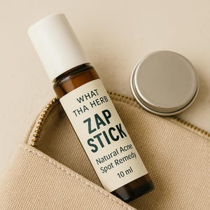 May include: A brown glass bottle with a white cap and label reading "ZAP STICK Natural Acne Spot Remedy 10 ml." A small silver container sits next to the bottle, both resting on a beige zippered pouch. The product is from "WHAT THA HERB."