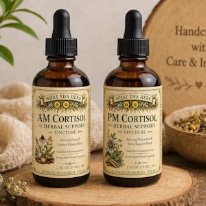 Può includere: Due flaconi di vetro ambrato di tintura a base di erbe con contagocce neri, etichettati "AM Cortisol" e "PM Cortisol". Le etichette presentano illustrazioni botaniche e il testo "What Tha Herb". Le bottiglie sono su una superficie di legno.