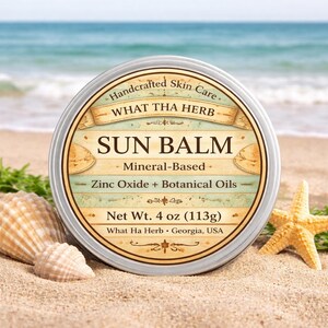 May include: A round, silver tin of "SUN BALM" with the text "Handcrafted Skin Care" and "WHAT THA HERB". The label also states "Mineral-Based", "Zinc Oxide + Botanical Oils", and "Net Wt. 4 oz (113g)". The tin is on a sandy beach with seashells and a starfish.