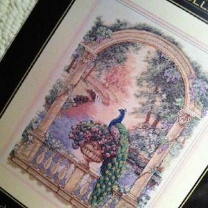 May include: A cross-stitch kit package titled The Gold Collection featuring a needlepoint design of a peacock perched on a stone balcony railing next to a large urn filled with pink flowers, framed by a stone archway with trees and foliage.