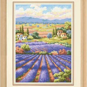 Puede incluir: Un bordado en punto de cruz enmarcado que representa un paisaje con hileras de campos de lavanda morada en primer plano, pequeñas casas de piedra, árboles verdes y colinas bajo un cielo azul con nubes suaves, en un marco de madera clara.