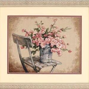 May include: A framed cross-stitch embroidery depicting a bouquet of pink roses arranged in a grey metal bucket, which is placed on a wooden chair. The artwork features a textured beige background and is enclosed in a decorative light-coloured frame.
