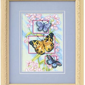 May include: A framed cross-stitch embroidery featuring three butterflies in shades of blue, yellow, and black, surrounded by pink flowers and green stems. The artwork is set against a light blue background and enclosed in a decorative cream-coloured frame.
