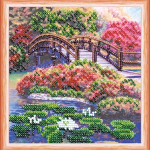 May include: A framed beaded art piece depicting a Japanese garden scene. A wooden arched bridge spans a pond with lily pads. Lush foliage in shades of green, red, and pink surrounds the bridge, creating a serene and colorful landscape.