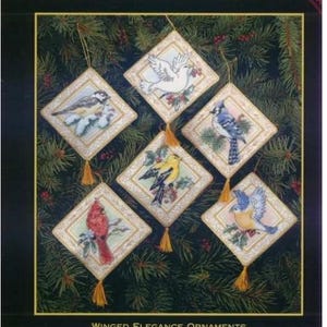 Op de afbeelding: Een telpatroon borduurpakket genaamd Winged Elegance Ornaments met zes ruitvormige ornamenten met illustraties van een witte duif, een blauwe gaai, een kardinaal en andere vogels op takken, elk met een gouden kwastje aan de onderkant.