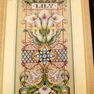 May include: A framed cross-stitch artwork displaying a floral design with the word "LILY" at the top. The piece features white calla lilies with green foliage, detailed patterns, and a neutral backdrop. It is presented in a light-coloured wooden frame.