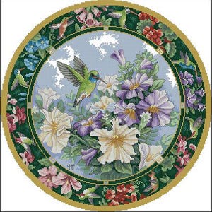 May include: A circular cross-stitch pattern featuring a green hummingbird hovering near white and purple petunia flowers against a light blue sky. The design is framed by a dark green border filled with smaller hummingbirds and floral motifs.
