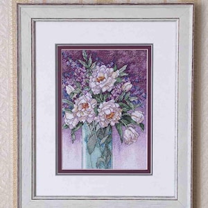 May include: A framed cross-stitch embroidery depicting a bouquet of white and light pink peonies with green leaves in a tall, translucent blue vase. The artwork is set against a deep purple background and enclosed in a white wooden frame.
