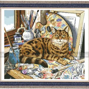 May include: A framed cross-stitch embroidery depicting a brown tabby cat resting in an artist's studio. The scene includes a wooden palette, a jar of brushes, tubes of paint, a glass jar with a brush, and an ink bottle, all set within a dark blue frame.