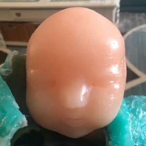 Op de afbeelding: Een gladde, perzikroze poppenkop met gesloten ogen en een kleine, gedefinieerde neus en mond. De kop rust op een turquoise oppervlak. De poppenkop is lichtroze.