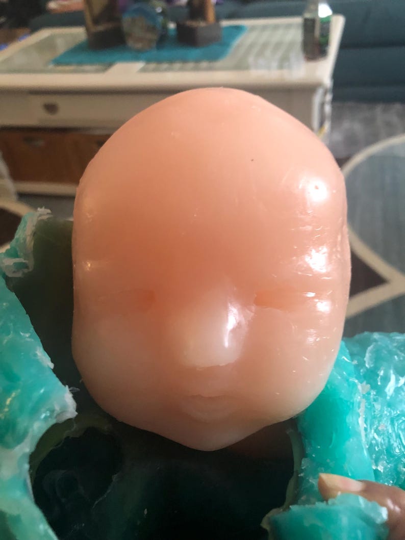 Op de afbeelding: Een gladde, perzikroze poppenkop met gesloten ogen en een kleine, gedefinieerde neus en mond. De kop rust op een turquoise oppervlak. De poppenkop is lichtroze.