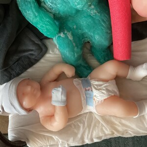 Op de afbeelding: Een levensechte babypop met een witte muts, wanten en sokken, liggend op een witte doek. De pop heeft een luier met een blauw en wit ontwerp. Een turquoise sculptuur en een rode zwemband zijn op de achtergrond.