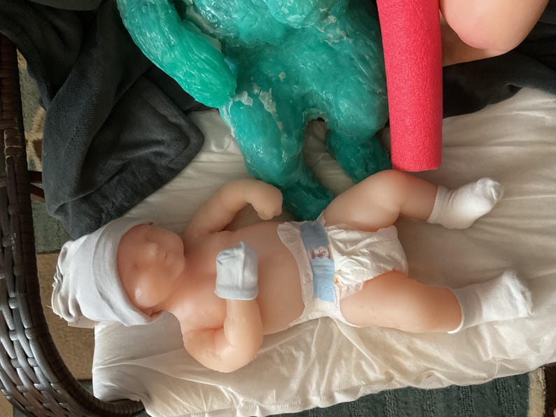 Op de afbeelding: Een levensechte babypop met een witte muts, wanten en sokken, liggend op een witte doek. De pop heeft een luier met een blauw en wit ontwerp. Een turquoise sculptuur en een rode zwemband zijn op de achtergrond.